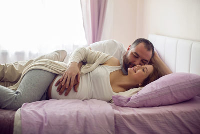 Husband in a white t-shirt with a pregnant wife sitting on the bed at home