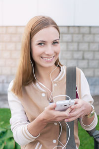 Portrait of smiling woman listening music while using mobile phone against wall