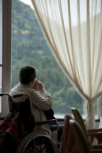 Rear view of man on wheelchair by window at home