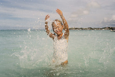 Cheerful girl splashing water in sea against cloudy sky
