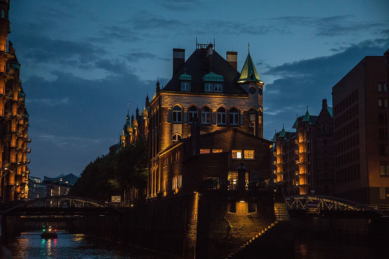 ILLUMINATED BRIDGE OVER RIVER AMIDST BUILDINGS IN CITY AT DUSK