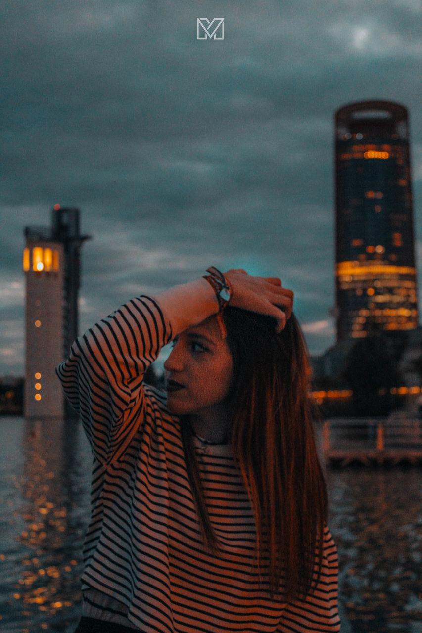 PORTRAIT OF YOUNG WOMAN STANDING BY ILLUMINATED CITY IN BACKGROUND