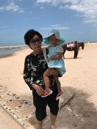 Portrait of grandmother holding granddaughter at beach against sky