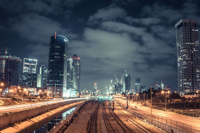 Illuminated railroad tracks in city against cloudy sky