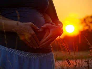 Cropped hands of man with pregnant girlfriend making heart shape on abdomen against orange sky