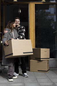 Smiling man embracing woman holding box while standing in front of apartment