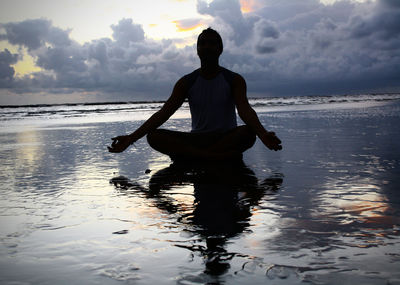 Silhouette man sitting in lotus position on shore at beach against sky