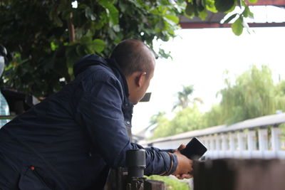 Side view of man holding mobile phone while leaning on railing