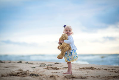 Cute girl holding stuffed toy while standing against sea at beach