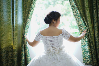 Rear view of bride in dress standing against window at home