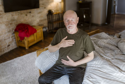 Senior man with eyes closed doing breathing exercise while sitting on bed at home