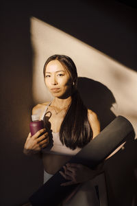 Portrait of young woman with exercise mat and water bottle standing against wall