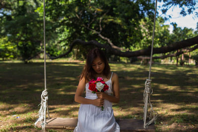 Young woman holding roses sitting on swing at land