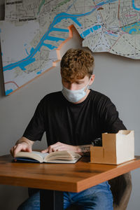 A young guy with curly hair with a medical mask on his face sits at a table in a cafe reading an exc