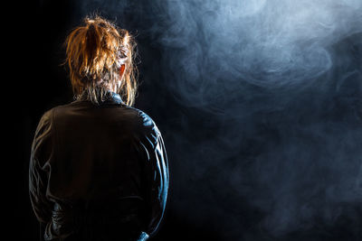 A woman with red hair and a leather jacket stands lonely in the dark