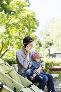 Mother using mobile phone while sitting with baby girl on bench at park