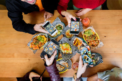 Top view crop anonymous friends taking pictures of delicious food on smartphones while gathering at table in restaurant