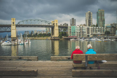 Rear view of senior couple sitting on bench by river in city