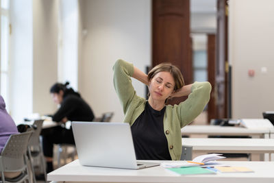 Tired female mature student taking break stretching arms while studying online in university library