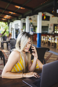 Focused young female in casual summer dress browsing netbook while talking on phone on outdoor restaurant terrace