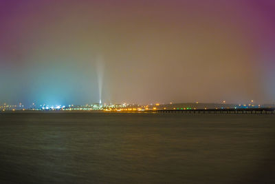 Illuminated cityscape by sea against sky at night