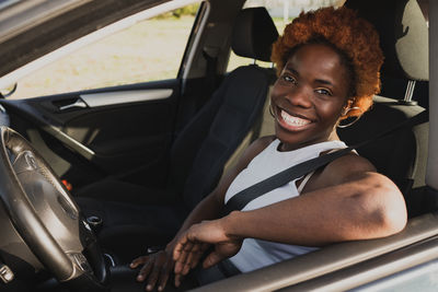 Side view of cheerful black female with afro hairstyle sitting on driver seat and looking at camera in sunny day