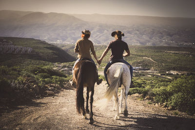 Rear view of couple holding hands while riding horses on field against mountain