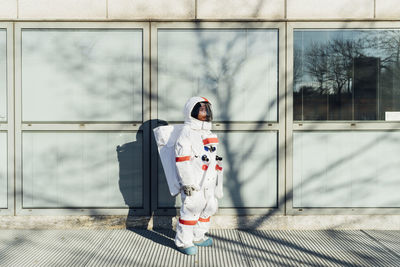 Young female astronaut in space suit standing by glass window in city during sunny day