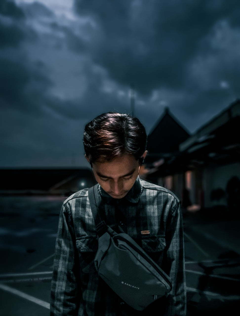 one person, darkness, cloud, sky, black, adult, young adult, architecture, men, night, standing, looking, front view, dark, waist up, focus on foreground, nature, casual clothing, city, screenshot, dusk, overcast, looking down, portrait, serious, outdoors