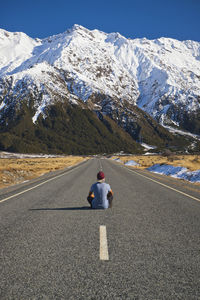 Rear view of man sitting on road against snowcapped mountain