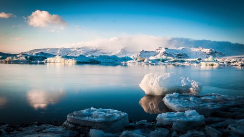 Scenic view of icebergs at lakeshore by snowcapped mountains against sky