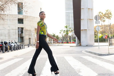 Side view full body of self assured young fashionable african lady with short hair in trendy outfit and high heels smiling and crossing road in city