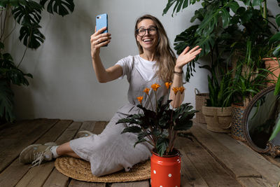 Full length of smiling woman taking selfie sitting by plants at home