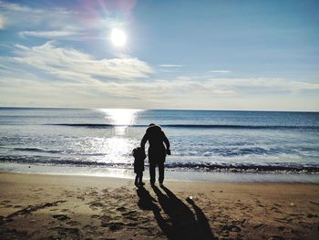 Mother and daughter walking at beach against sky during sunset