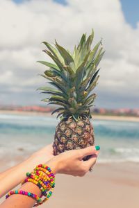 Hands of woman holding pineapple at beach against sky