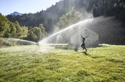 Woman jumping on grass near sprinkler during sunny day