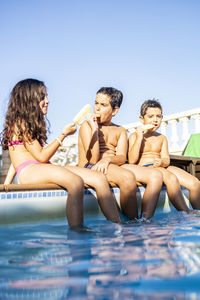 Friends eating popsicle while sitting on poolside against clear sky