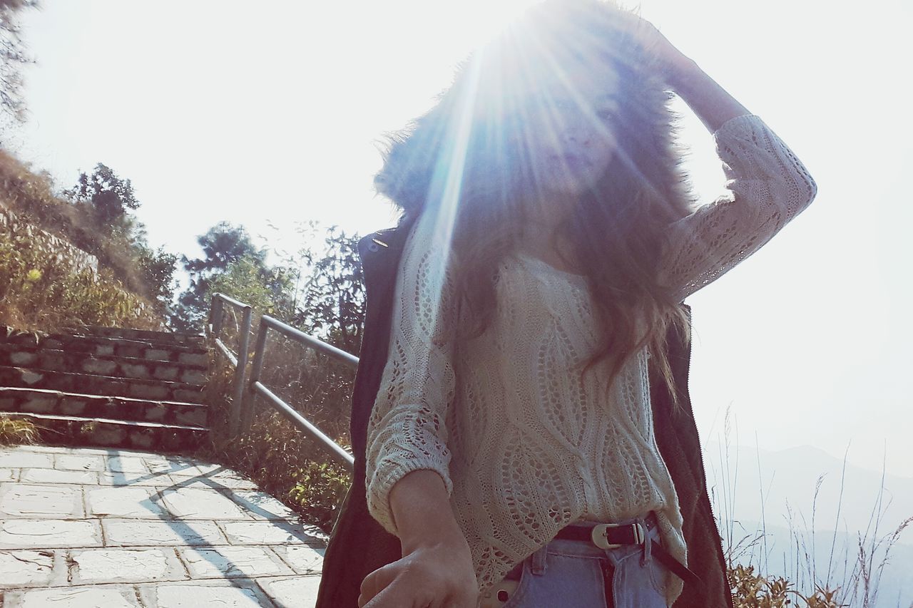 lifestyles, sunlight, sunbeam, leisure activity, young adult, standing, tree, clear sky, young women, casual clothing, sun, lens flare, long hair, day, sunny, built structure, three quarter length, person