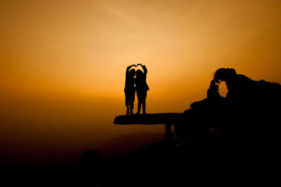 Silhouette people making heart shape on cliff against sky during sunset