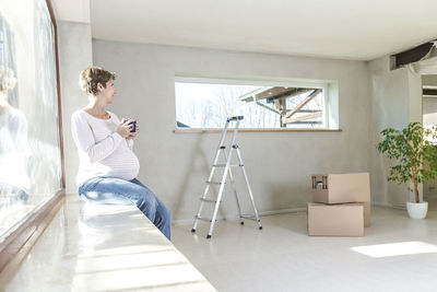 Pregnant woman holding cup while sitting by window at home