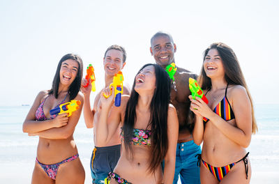 Portrait of smiling friends holding squirt guns while standing at beach against sky