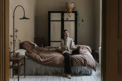 Happy non-binary person talking on smart phone while sitting in bedroom at home