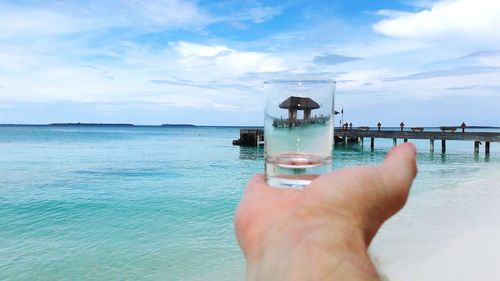 Cropped hand of person holding shot glass against sea and sky