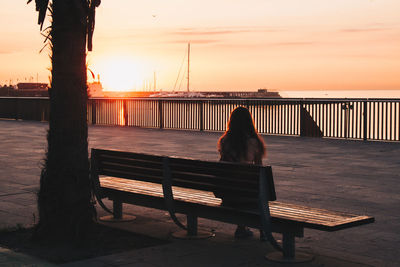 Woman sitting on bench looking at sea against sky during sunset