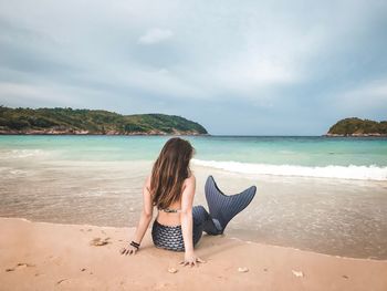 Rear view of woman sitting at beach against cloudy sky