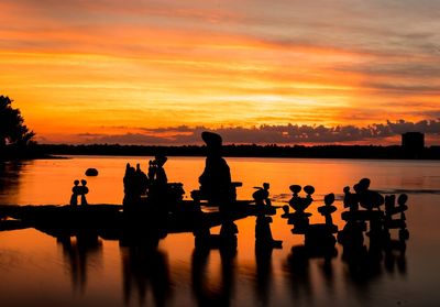 Silhouette people sitting on lake against orange sky