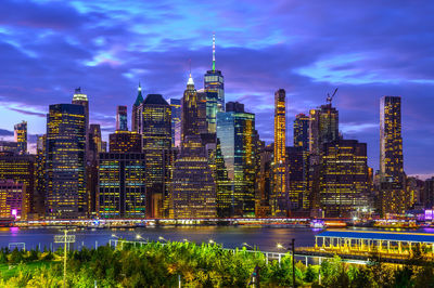 Illuminated modern buildings by river in city against sky at night