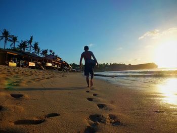 Rear view of man walking on beach against sky during sunset