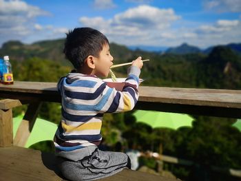 Side view of boy eating food while sitting on bench against mountains