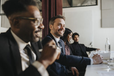 Smiling businessman looking away while sitting with colleagues during meeting at office